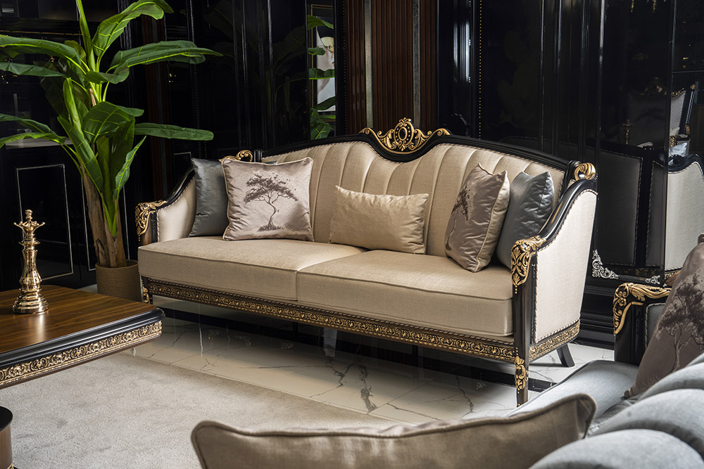 Elegant beige sofa with gold filigree detailing in a black, glossy-walled living room, surrounded by plush cushions and a tall plant.