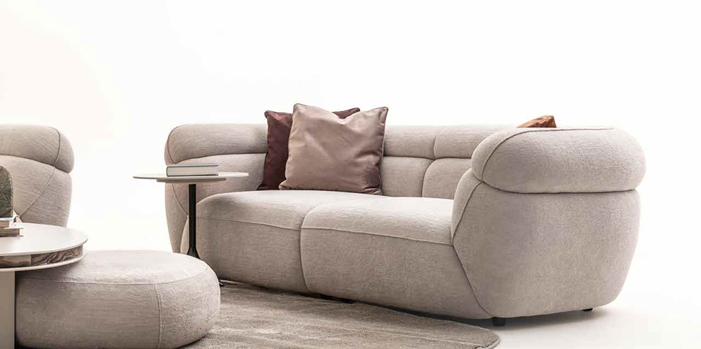 Beige modular sofa with rounded arms and two decorative pillows, plus a round ottoman and small side table in a minimalist living room setting.