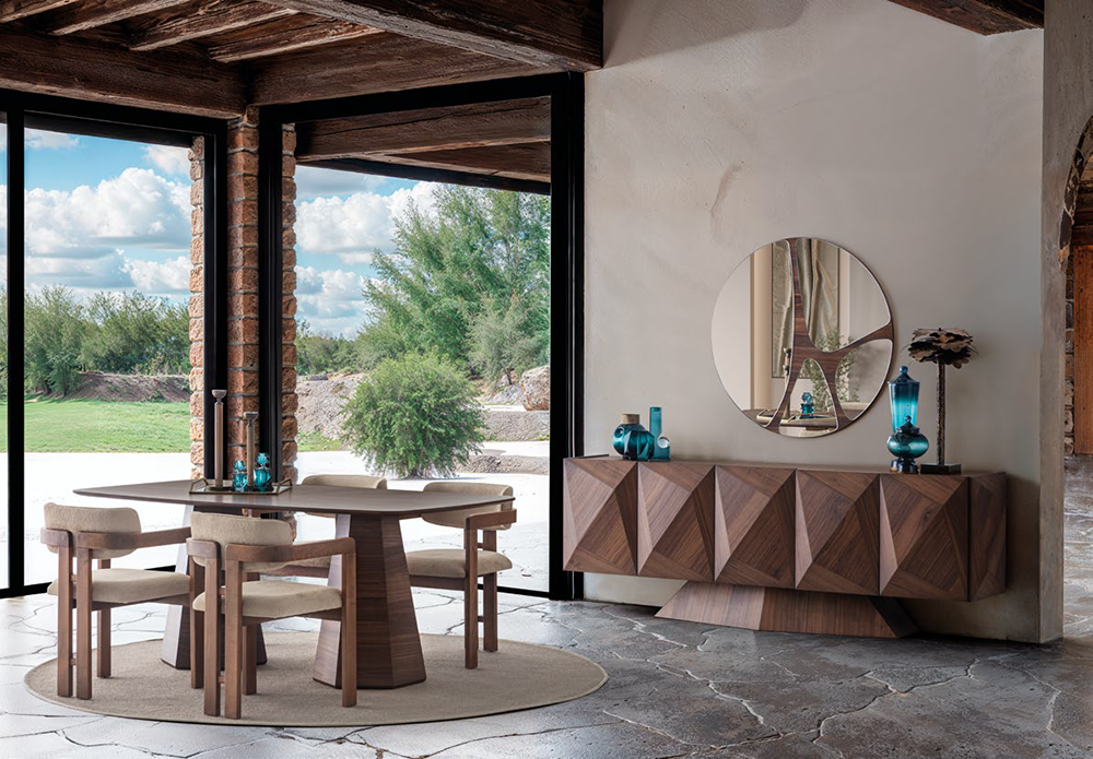 Modern dining room with a round wooden table and six upholstered chairs, large glass doors, and a geometric-patterned sideboard with blue decor.