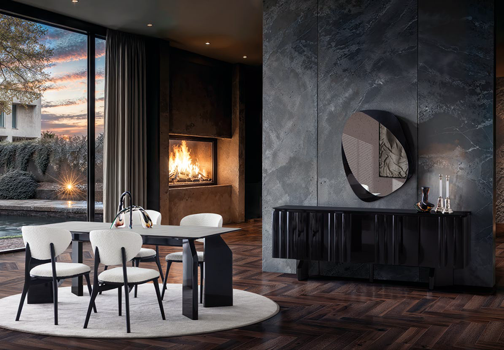 Modern dining area with white upholstered chairs around a rectangular table, a lit fireplace, and a large window showing a sunset outside.