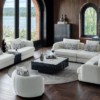 Luxurious living room with white sectional sofas arranged around a black coffee table on a circular rug fireplace to the right and large arched windows revealing a lakeside view