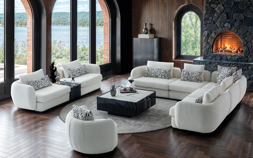 Luxurious living room with white sectional sofas arranged around a black coffee table on a circular rug, fireplace to the right, and large arched windows revealing a lakeside view.