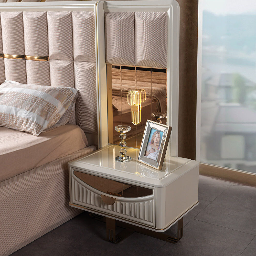 White nightstand with glass top and gold trim beside a tufted bed, featuring a framed photo and a gold lamp.