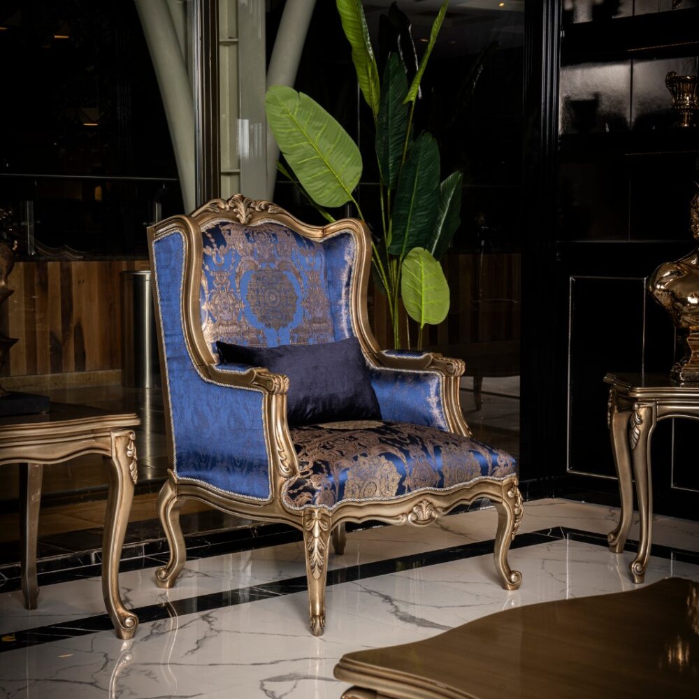 Elegant blue and gold damask armchair with carved frame in a luxe indoor setting, next to a tall green plant and marble floor.