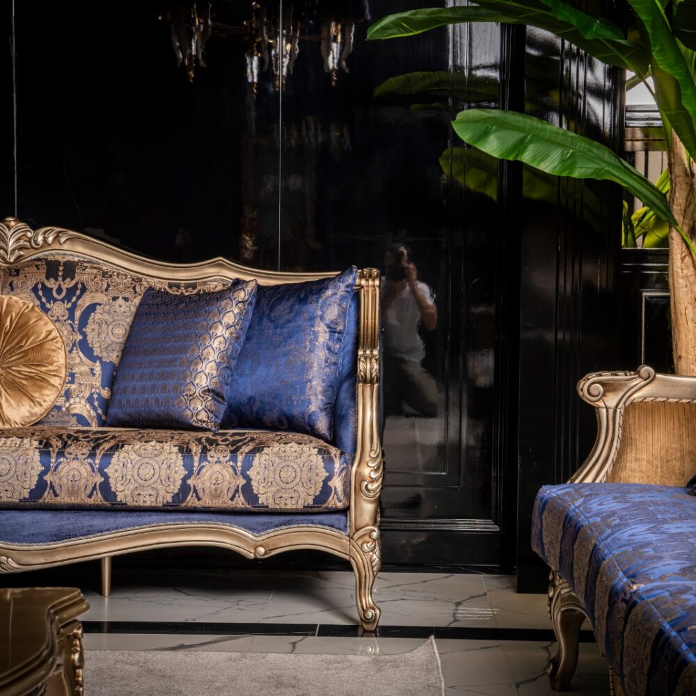 Luxurious blue and gold damask sofa with a round gold cushion, in an opulent living room with dark walls and green plants.
