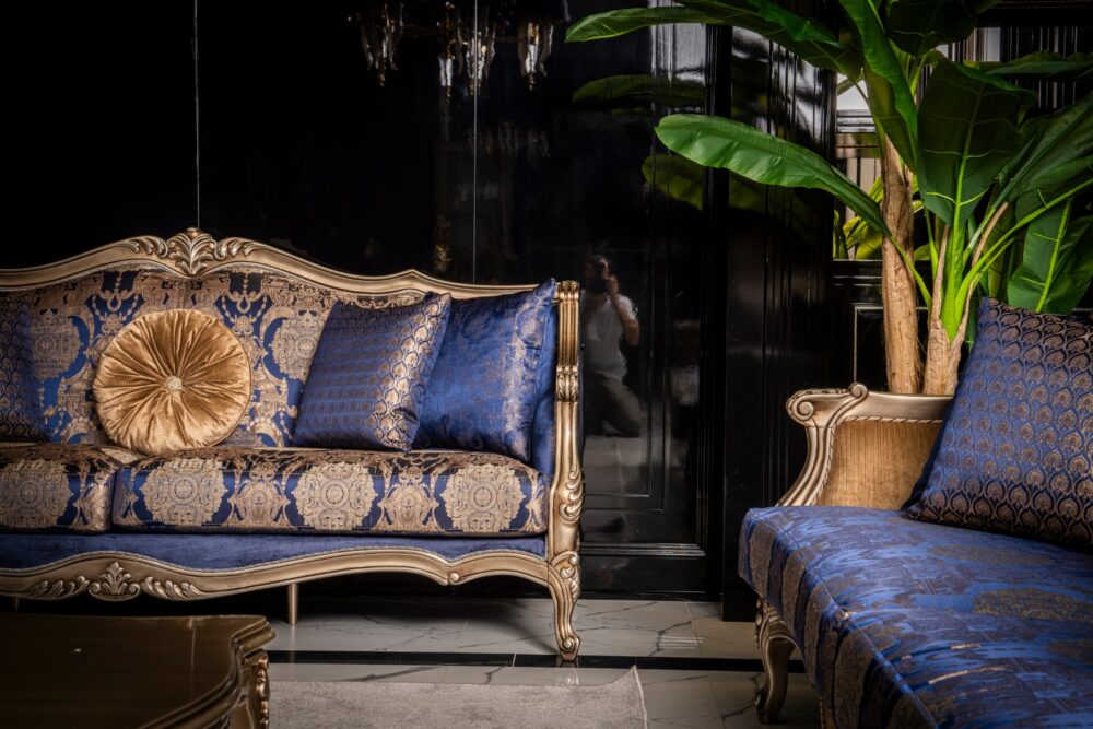 Emirgan Classic Set 39 Armonna Luxurious blue and gold damask sofa with a round gold cushion in an opulent living room with dark walls and green plants