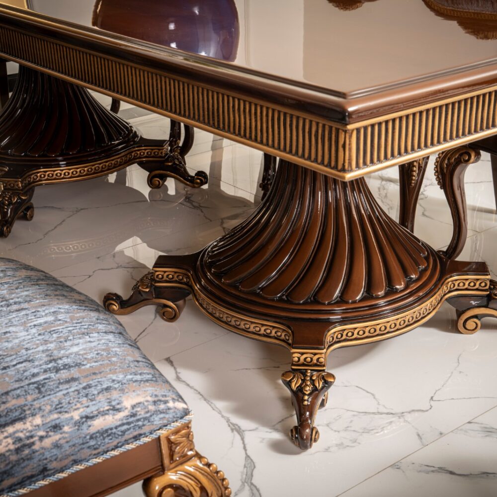 Close-up of an ornately carved dining table pedestal with fluted, dark wood base and gold trim on a marble floor.
