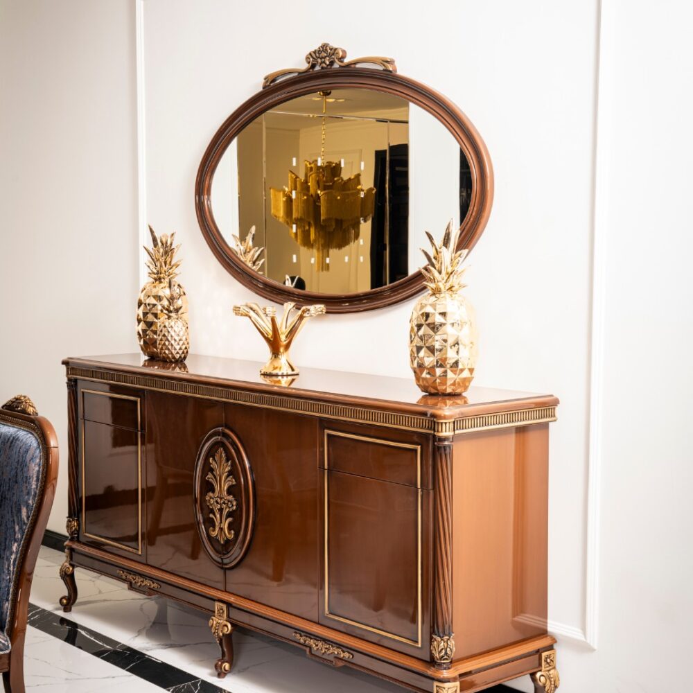 Elegant wooden sideboard with gold trim and an oval mirror above, topped with gold pineapple decor and a gold centerpiece on a marble floor.