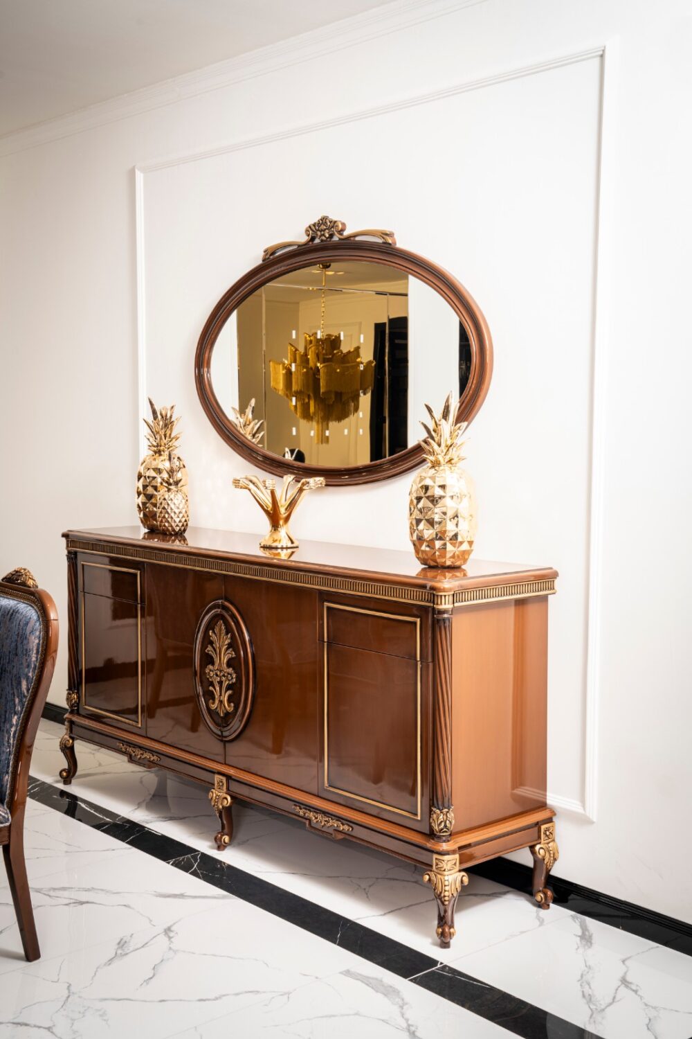 Gize Classic Set 2 Armonna Elegant wooden sideboard with gold trim and an oval mirror above topped with gold pineapple decor and a gold centerpiece on a marble floor