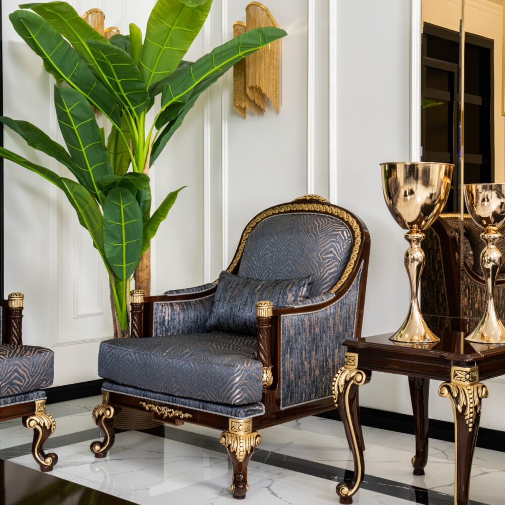 Luxurious living room corner with a tall green plant, blue patterned chair, and gold-accented furniture.