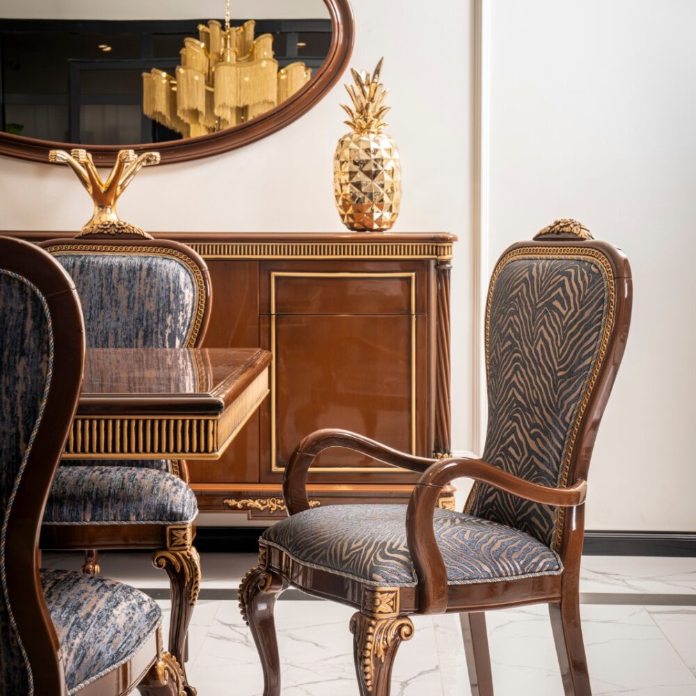 Elegant antique wood dining set with blue patterned upholstery and gold accents against a light wall. A gold pineapple decor and a large oval mirror reflect a chandelier in the background.