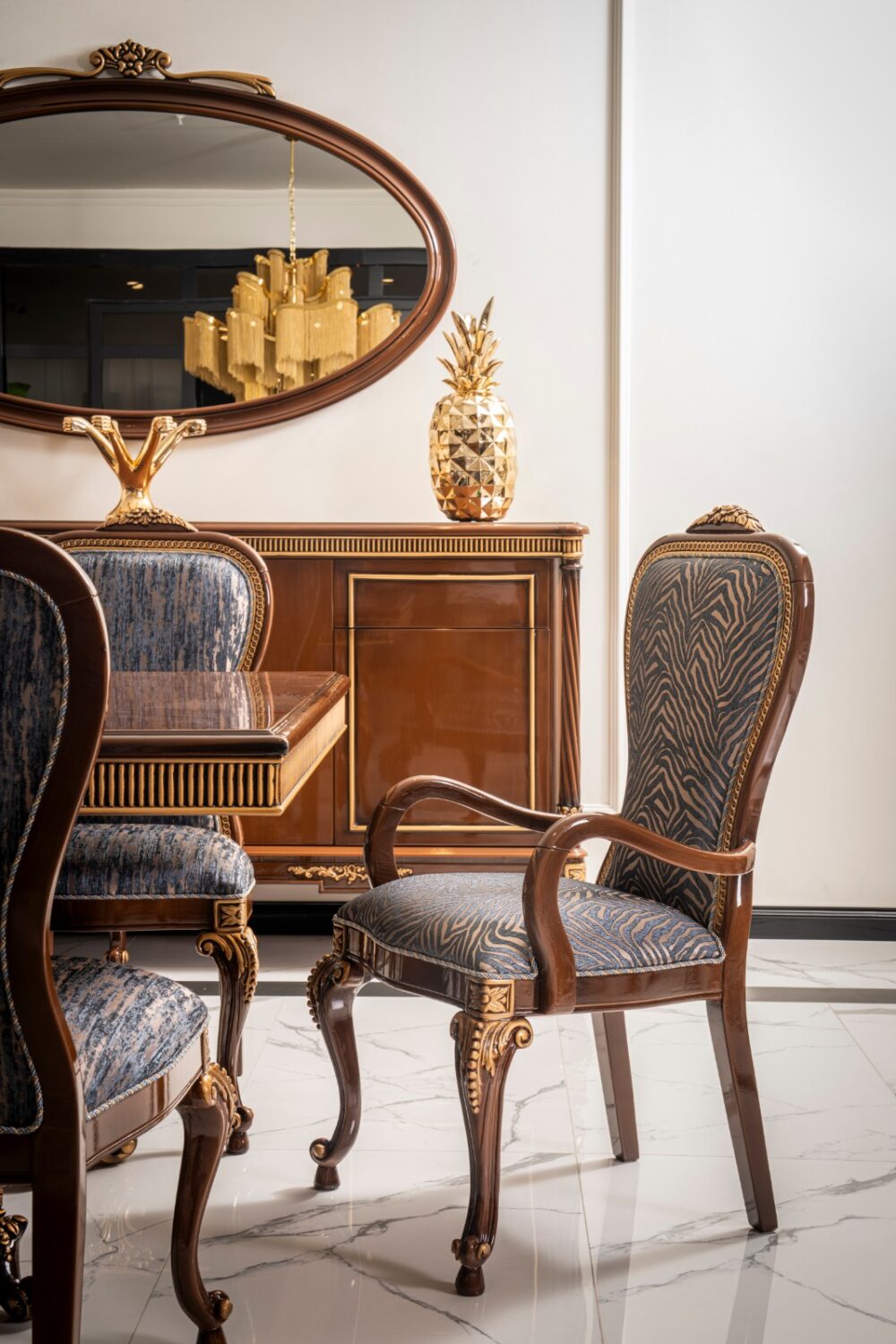 Gize Classic Set 7 Armonna Elegant antique wood dining set with blue patterned upholstery and gold accents against a light wall A gold pineapple decor and a large oval mirror reflect a chandelier in the background