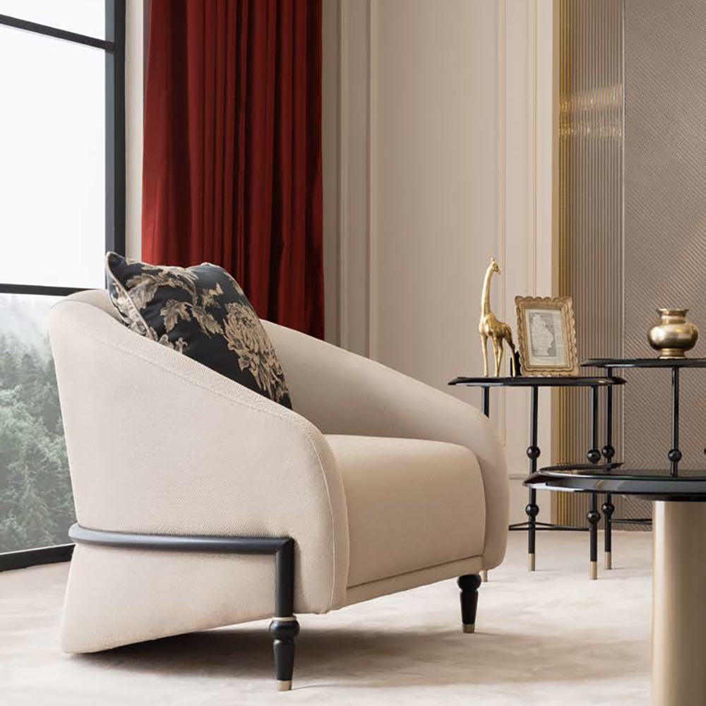 Beige upholstered armchair with a dark wooden leg, beige carpet, and a side table displaying a gold giraffe statue and framed photo by red velvet curtains.