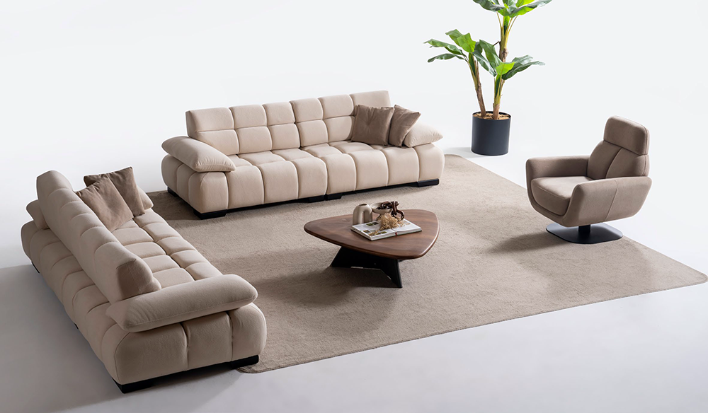 Beige modular sofa set with matching armchair on a large cream rug, wooden coffee table, and a potted plant in a bright showroom.