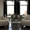 Lora Living Room 2 Armonna Two beige upholstered armchairs face a small round coffee table in a modern living dining space with large windows and potted plants