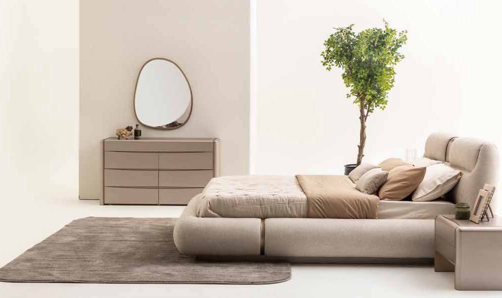 Modern neutral bedroom with a low upholstered bed, beige bedding, and a gray rug; dresser with a rounded mirror and a tall plant nearby.