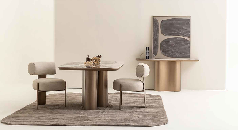 Minimalist dining area with a round marble-top table and two beige upholstered chairs on a textured rug, against a light backdrop, with abstract art on a pedestal nearby.