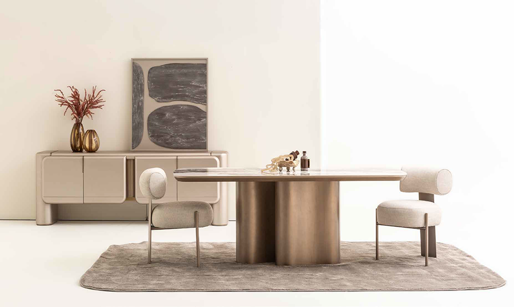 Modern dining room with a round table on a brass cylindrical base, two upholstered chairs, and a beige rug beneath.