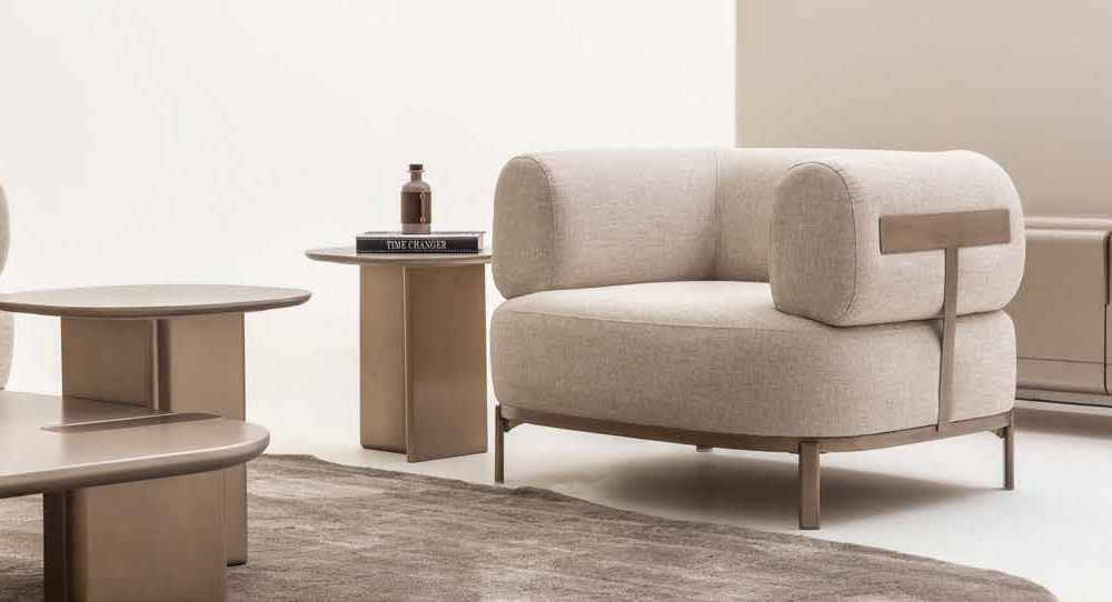 Beige upholstered lounge chair with curved arms in a neutral living room, beside a round coffee table and a side table with a bottle and book.
