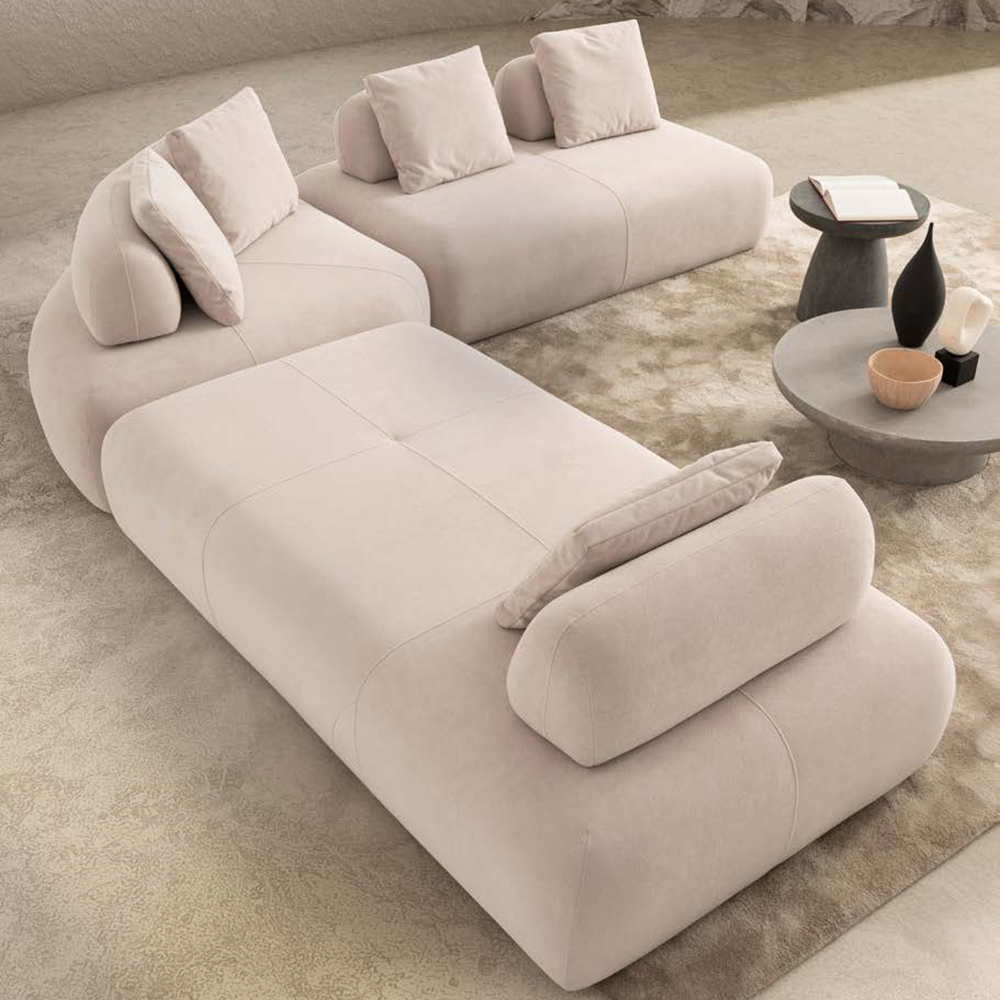Beige modular sofa with chaise and cushions in a neutral living room, beside round coffee tables with books and decorative vases.