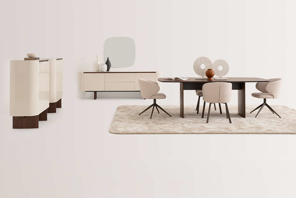 Modern dining room scene with a dark wood rectangular table surrounded by beige upholstered chairs on a light rug, and a light sideboard with decorative objects against a pale wall.