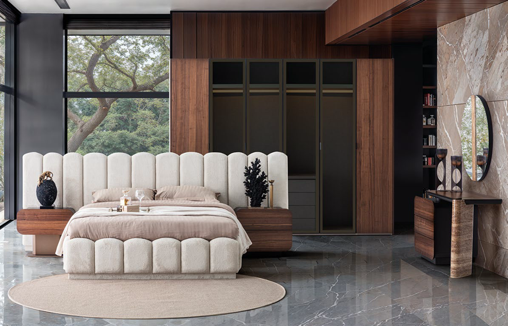 Luxurious modern bedroom with a plush beige upholstered bed, circular rug, wood accents, and floor-to-ceiling windows overlooking trees.
