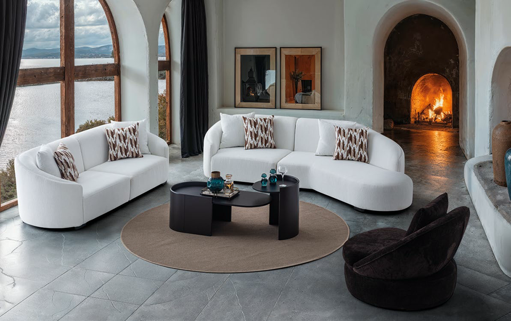 Bright modern living room with two white curved sofas around a black oval coffee table on a circular beige rug by a lit fireplace.