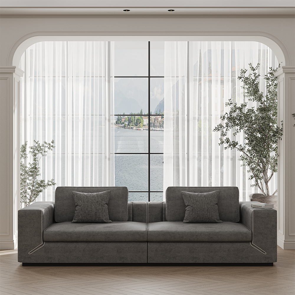 Gray sectional sofa with two cushions facing a large window overlooking a lake or coast, framed by sheer white curtains and tall plants in a light-filled living room
