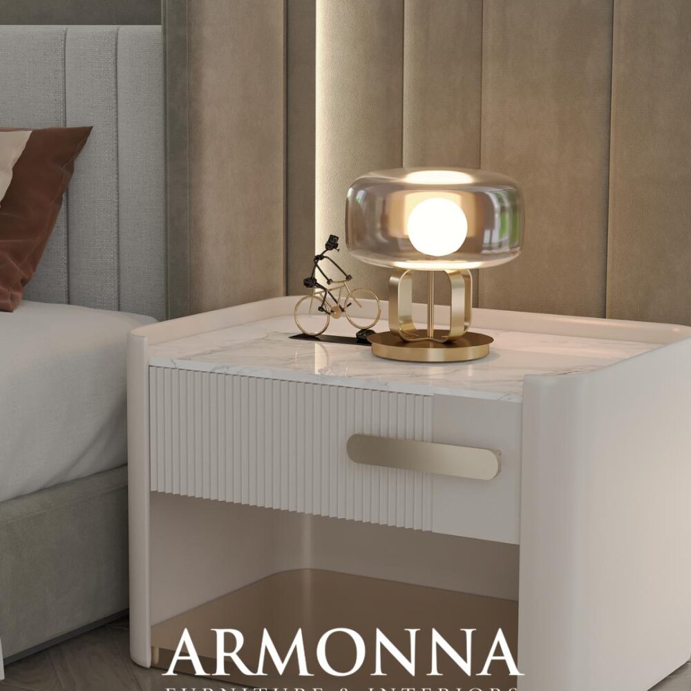 Modern white bedside table with a marble top, a gold lamp, and a small decorative bicycle on a softly lit bedroom surface (brand: ARMONNA Furniture & Interiors).