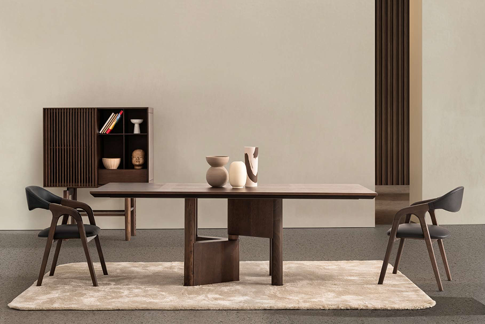 A modern wooden dining table with two matching chairs in a minimalist room, beige rug, and a shelf with ceramics in the background.