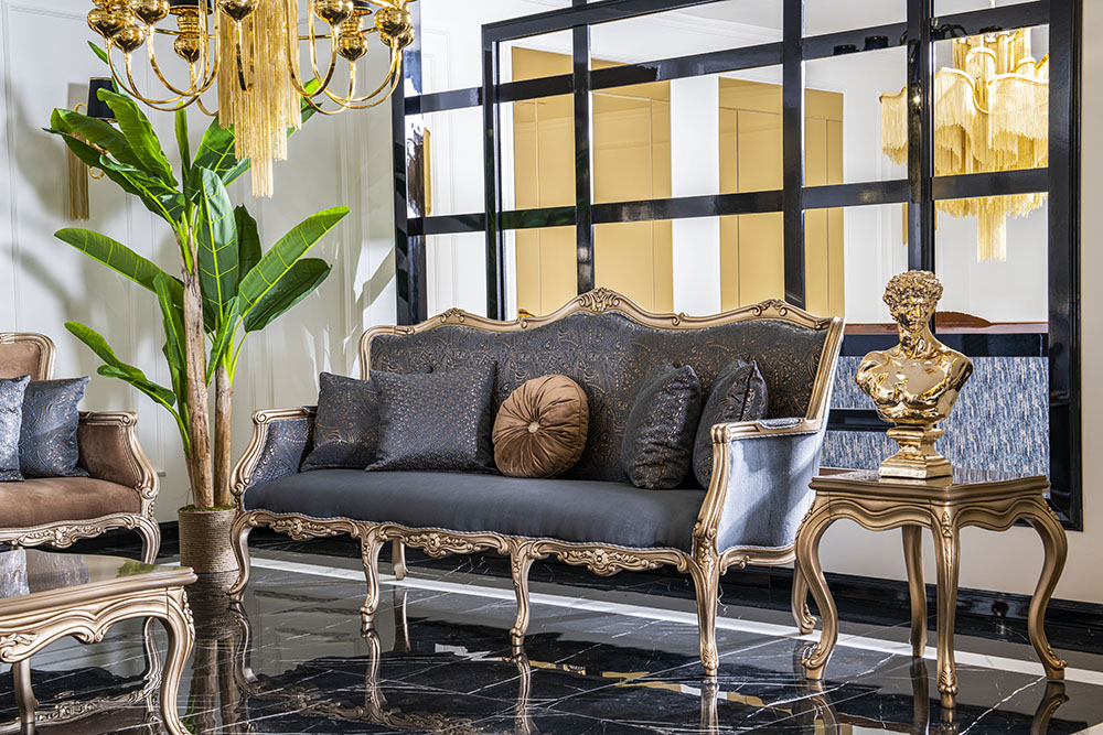 Luxurious living room with ornate gold-trimmed sofa, matching chairs, and dark throw pillows under a chandelier.