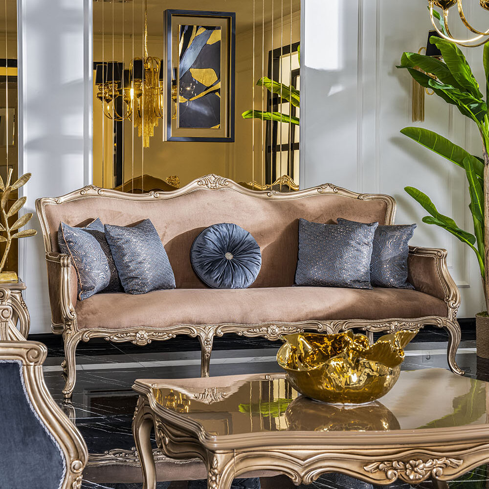 Elegant living room with a gold-framed sofa, matching ornate coffee table, blue-gray cushions, and a gold bowl centerpiece, set on a black-and-white tiled floor with chandeliers and a large green plant.