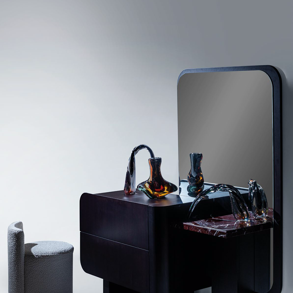 Dark modern vanity with rounded edges and a tall mirror, topped by colorful glass sculptures; a textured gray stool sits to the left.
