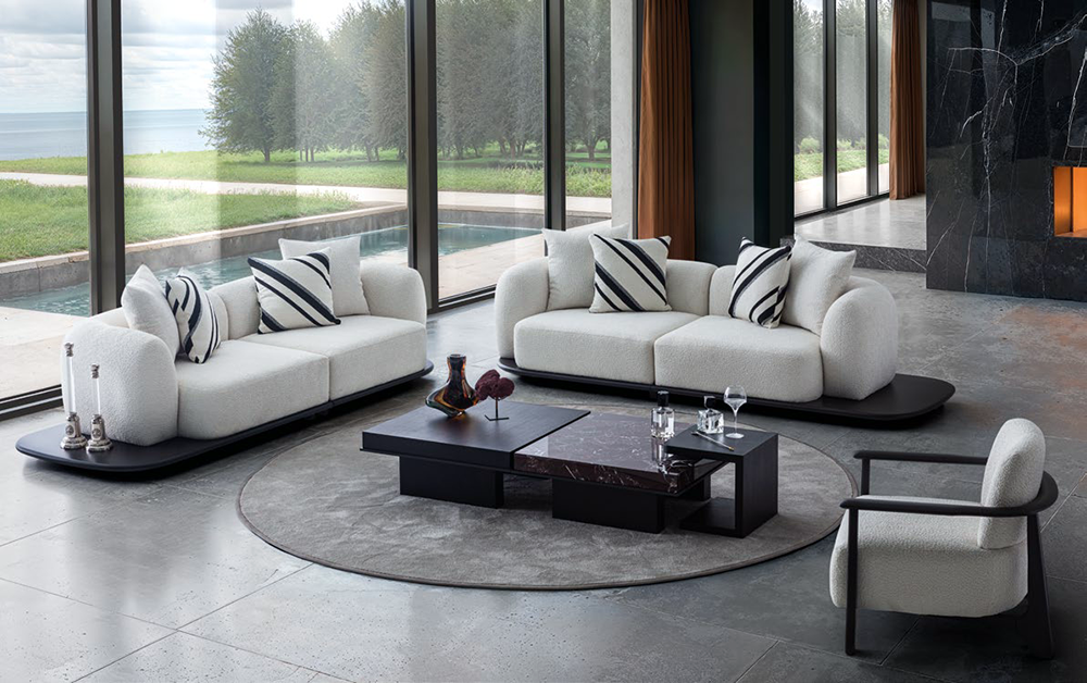 Modern living room with white curved sofas, a dark coffee table, and a circular rug by floor-to-ceiling windows showing an outdoor view of trees and water.
