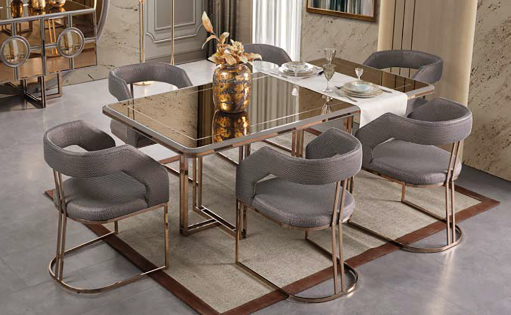Elegant dining room with a glass-top table and six gray upholstered chairs with gold metal legs, centerpiece vase, and place settings.