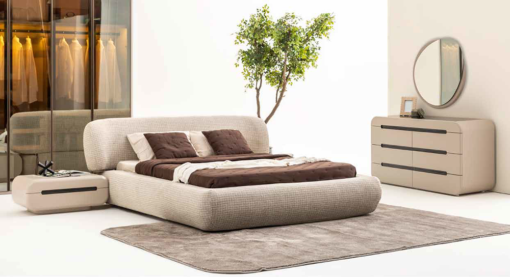 Modern beige upholstered bed with brown bedding and pillows in a minimalist bedroom with a rug and a dresser with round mirror on the right.