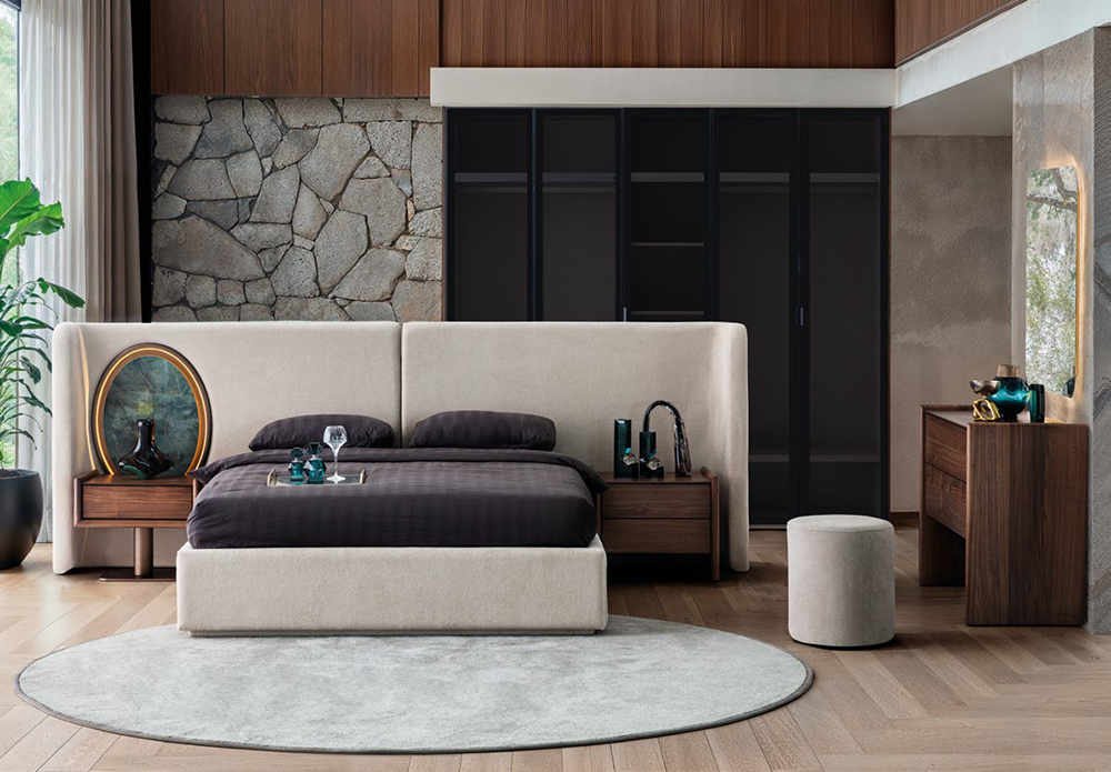 Luxurious modern bedroom with a beige upholstered platform bed, dark bedding, and wood nightstands against a stone accent wall.