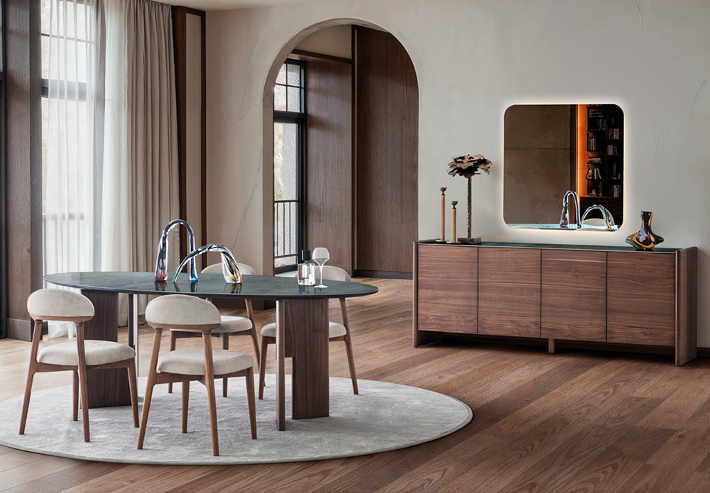 Round dining table with four cushioned chairs on a light rug in a modern dining area with wood floors and large windows with sheer curtains.