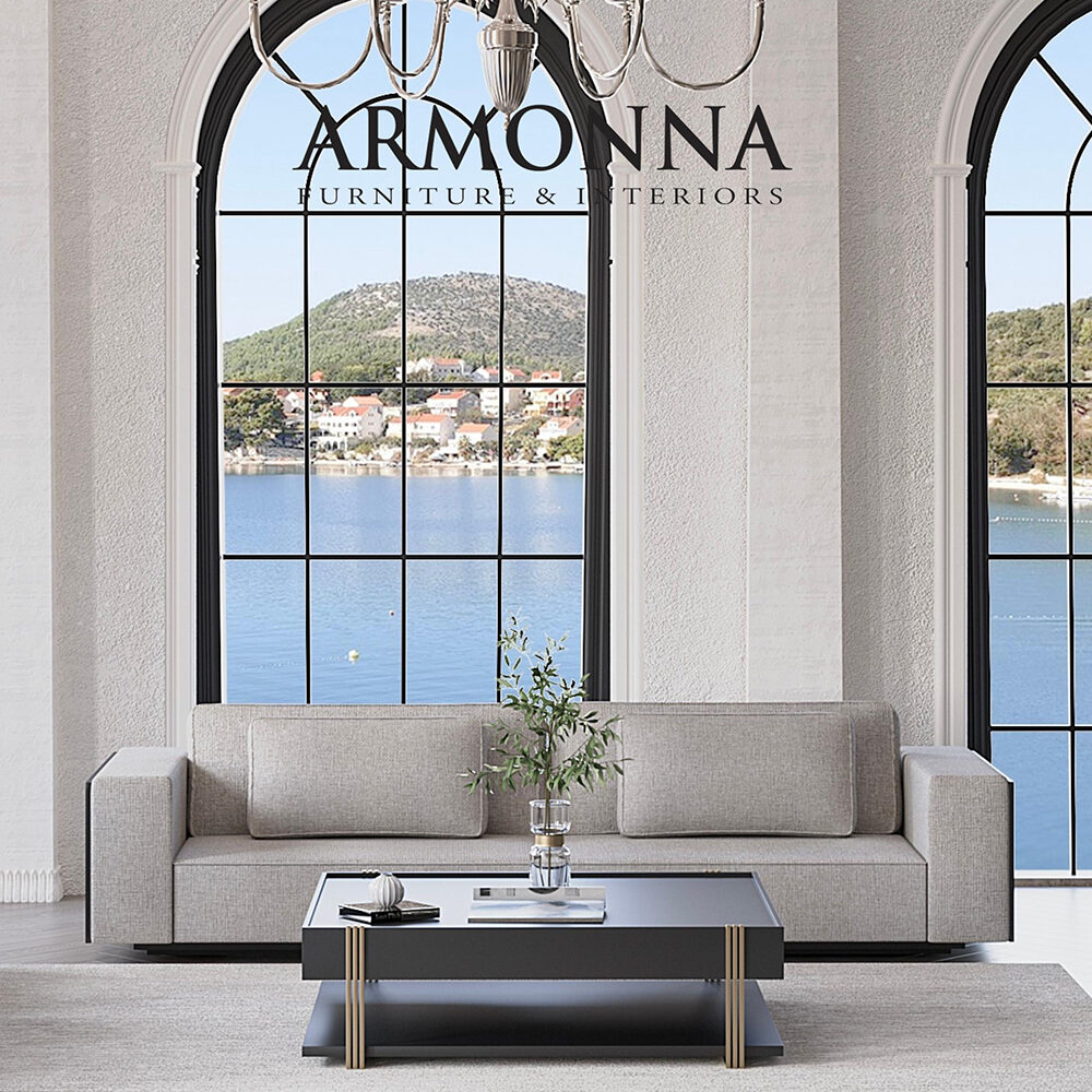 Luxurious living room with a beige sofa, glass coffee table, and crystal chandelier, framed by large arched windows showing a lake and hills; Armonna Furniture & Interiors logo on the glass.