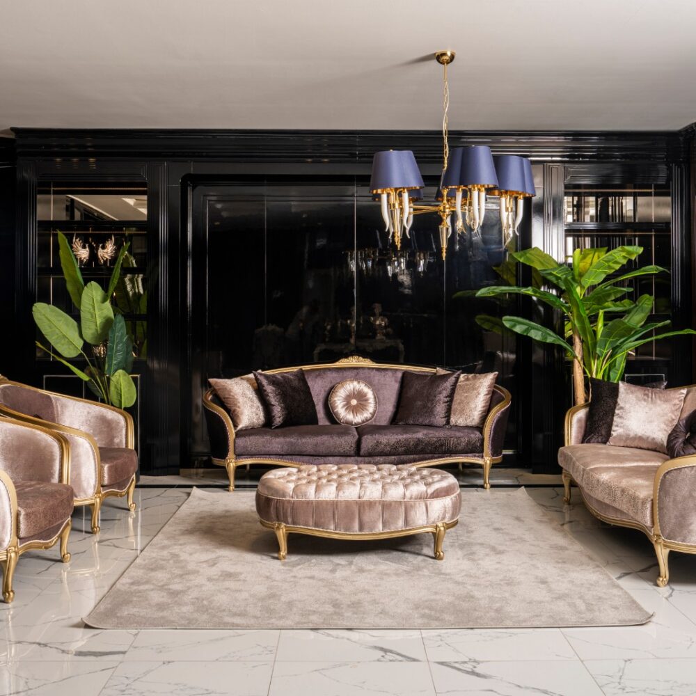 Luxurious living room with mauve velvet sofas, gold trim, and a tufted round ottoman under a blue-shaded chandelier.