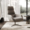 Modern gray swivel lounge chair with high back in a sunlit living room next to a glass topped table and sheer curtained windows overlooking a garden pond
