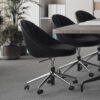Three black swivel chairs surround a wooden conference table in a bright meeting room with a potted plant and shelves in the background