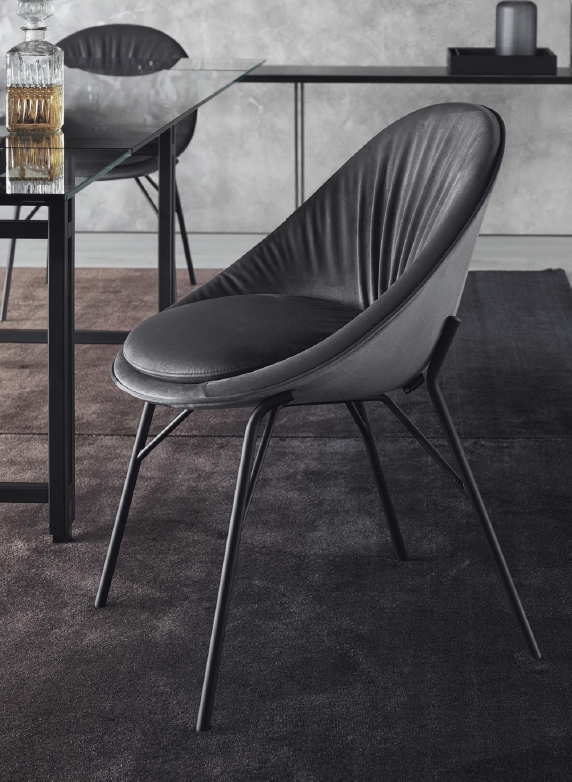 Gray curved lounge chair with slender metal legs beside a glass table in a modern office setting on a dark rug.