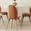 Four orange and beige dining chairs around a white round table with a fruit bowl in a bright minimalist dining room