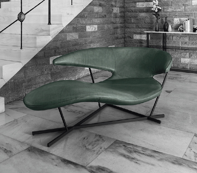 Green leather lounge chair with a metal X-base in a modern living area beside a brick wall and staircase.