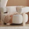 Cream boucle armchair with rounded arms and a warm orange brown throw draped over it in a sunlit living room setting