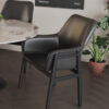 Black modern armchair with curved back and armrests beside a round marble table in a contemporary office space with potted plants in the background