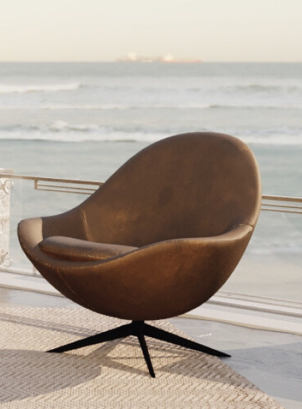 Brown egg-shaped lounge chair on a balcony with the ocean in the background. A modern swivel chair on a patterned outdoor rug.