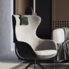 Modern beige lounge armchair with a black base and a dark throw pillow set in a contemporary bedroom with a bed and neutral decor