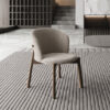 Beige upholstered lounge chair with wooden legs on a striped rug in a modern neutral toned living space with concrete floor and minimalist shelving in the background